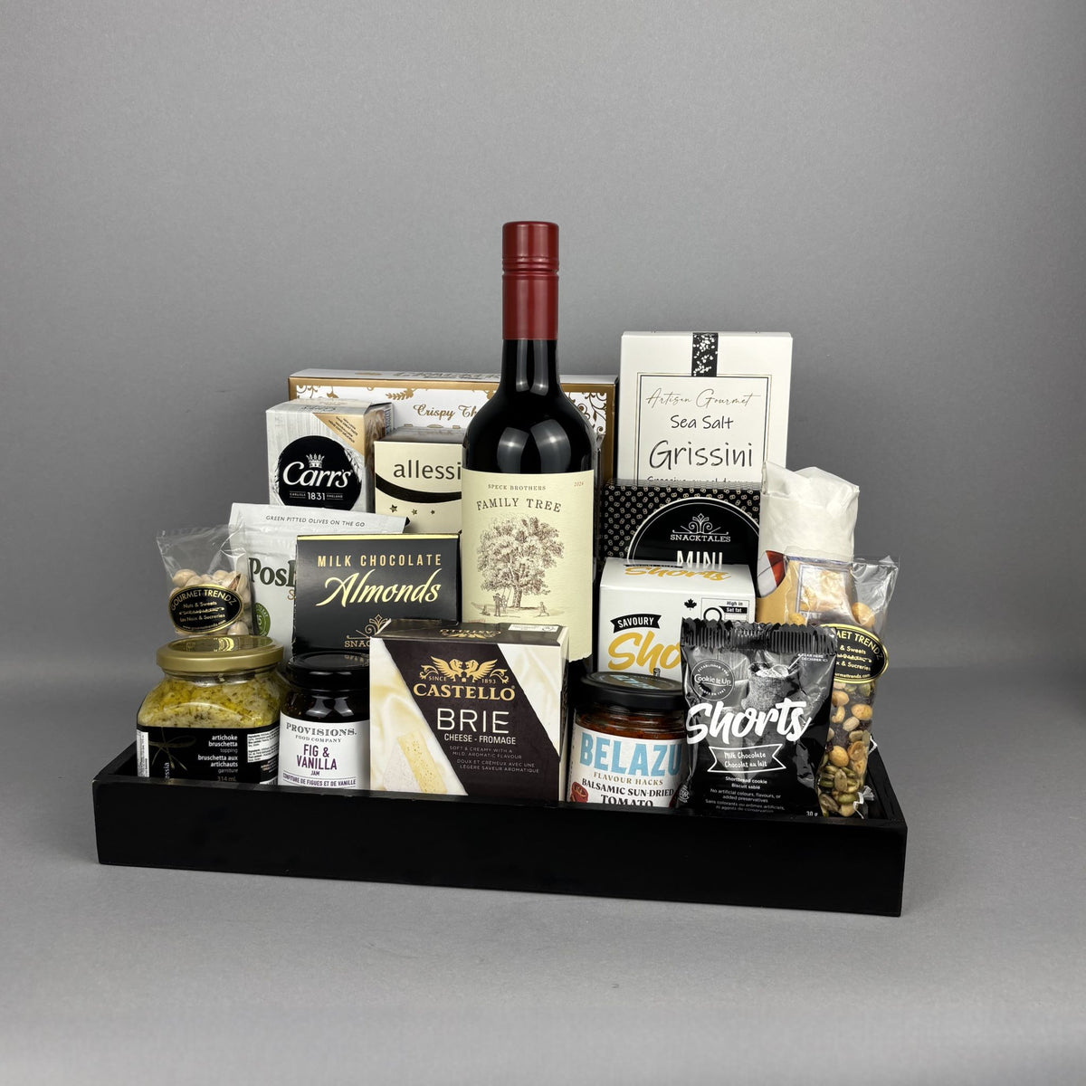 A gourmet gift hamper arranged in a black tray, featuring a bottle of Family Tree red wine, Castello Brie cheese, Belazu sauce, Sea Salt Grissini, Allessia biscotti, crackers, milk chocolate almonds, Provisions jam, and other packaged snacks and treats.