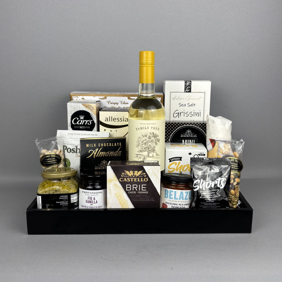 A gourmet gift hamper arranged in a black rectangular tray. The tray contains a bottle of white wine, assorted cheeses, chocolates, nuts, and packaged snacks.