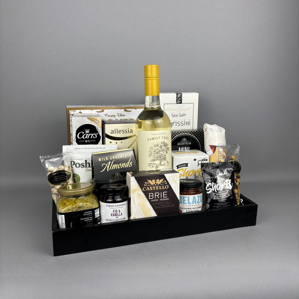 A gourmet gift hamper arranged in a black tray, featuring a bottle of white wine, cheese, crackers, nuts, and packaged snacks. Featuring brand names including Castello, Provisions, Belazu, Cookie It Up, and more.