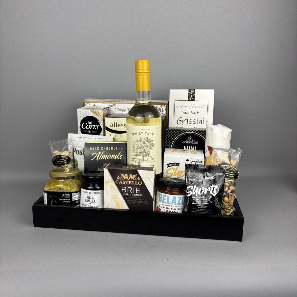 A gourmet gift hamper arranged in a black tray, featuring a bottle of Family Tree white wine, Carr's crackers and Allessia boxed biscotti, Castello Brie cheese, Belazu balsamic tomato spread, Canadian cheddar biscuits, and various other snacks.