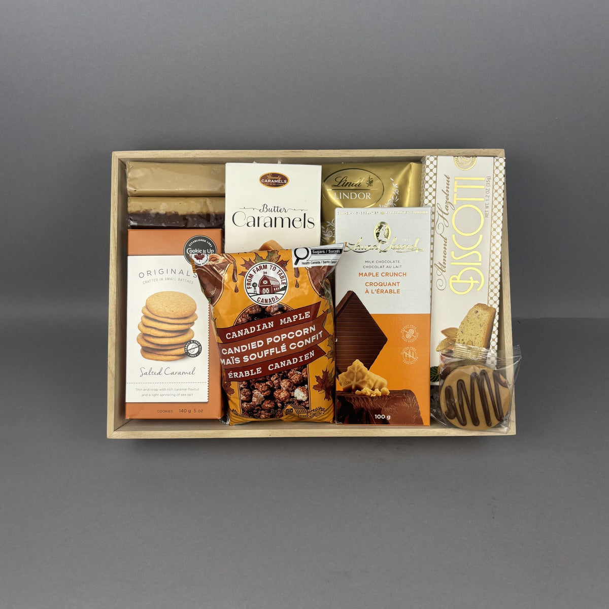 A gourmet gift box filled with assorted sweet treats, including Butter Caramels, Canadian Popcorn, Maple Fudge, and a Biscotti, presented in a light wood tray.