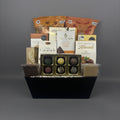 A gourmet chocolate gift basket arranged in a black rectangular basket. The basket contains assorted packaged chocolates, caramel candies, milk chocolate almonds, and decadent fudge.