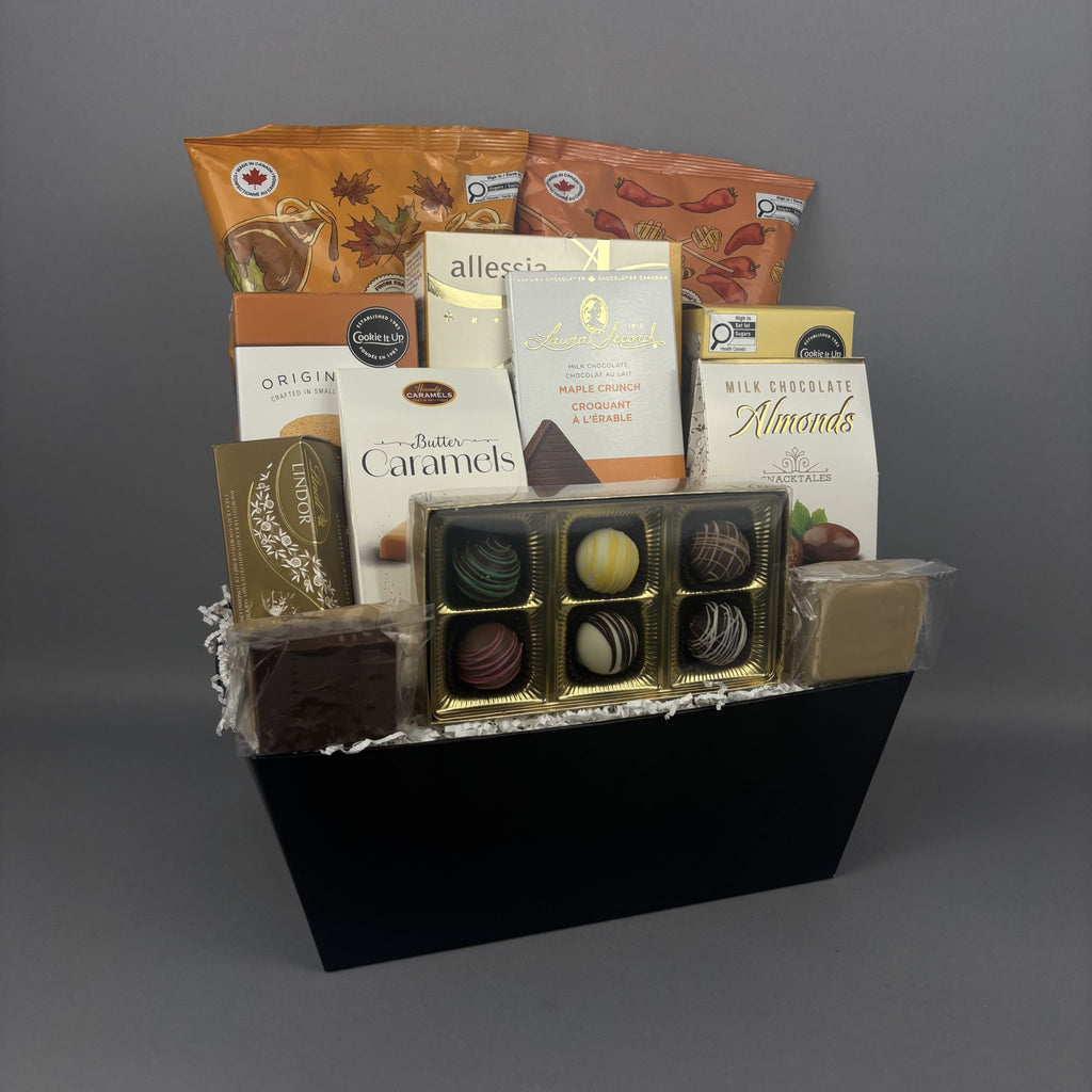 A gourmet chocolate gift basket arranged in a black rectangular box. The basket contains assorted packaged chocolates, milk chocolate bars, caramel candies, and a gold-trimmed box of assorted truffles, with branded packages including Alessia, Laura Secord, and Lindt.