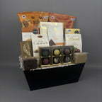 A gourmet chocolate gift basket arranged in a black rectangular box. The basket contains assorted packaged chocolates, milk chocolate bars, caramel candies, and a gold-trimmed box of assorted truffles, with branded packages including Alessia, Laura Secord, and Lindt.