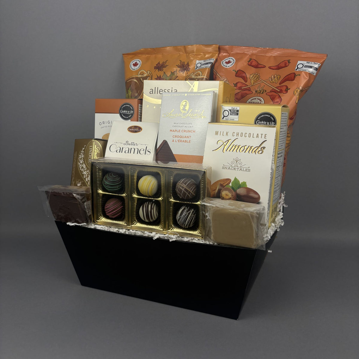A gourmet chocolate gift basket arranged in a black rectangular box. The basket contains assorted chocolates, including a box of gourmet handcrafted truffles, milk chocolate almonds, caramel candies, and packaged sweets from brands like Cookie It Up and Lindt.
