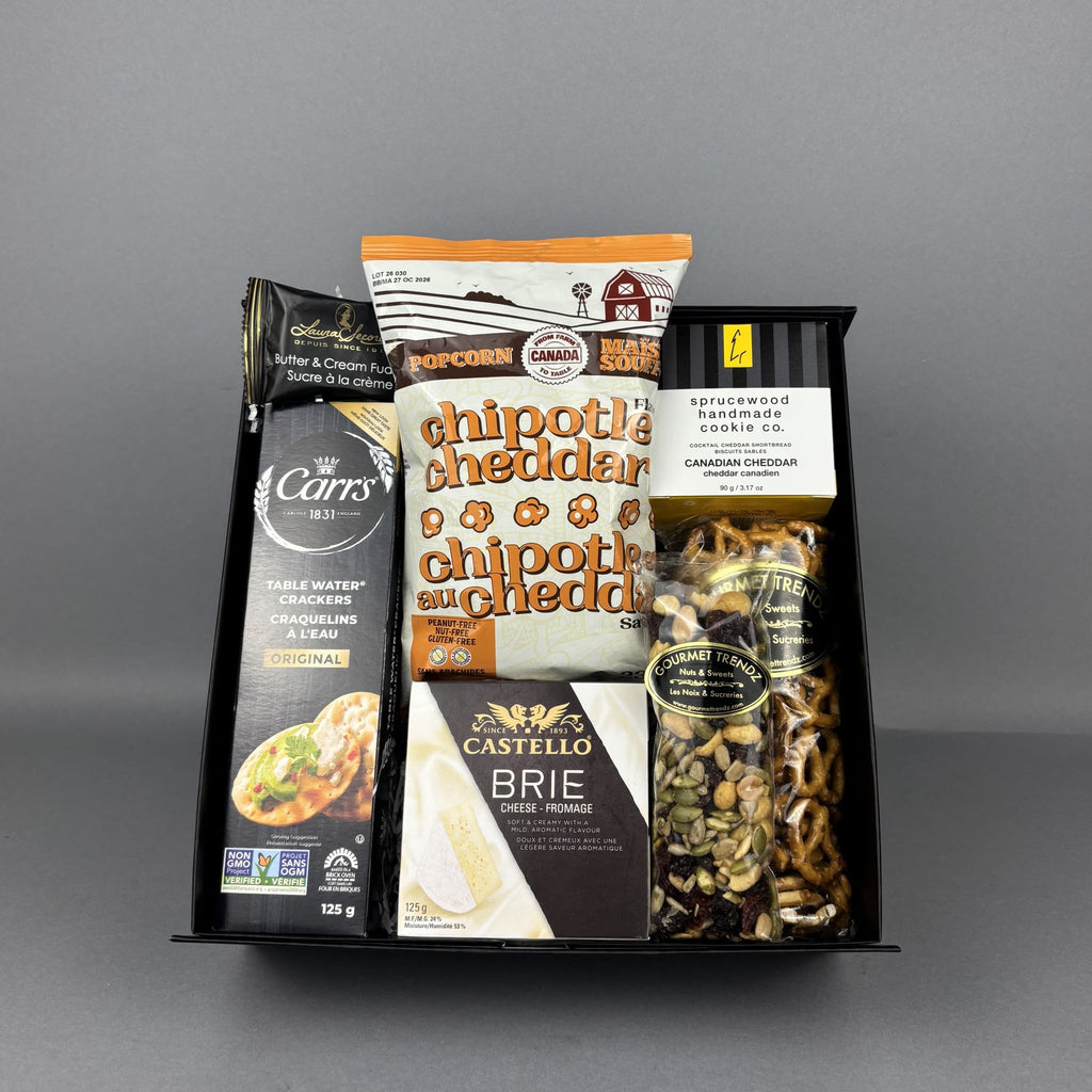 A curated snack and cheese gift basket arranged in a black box, featuring Carr's crackers, Castello Brie cheese, a bag of chipotle cheddar popcorn, and a bag of mixed nuts.