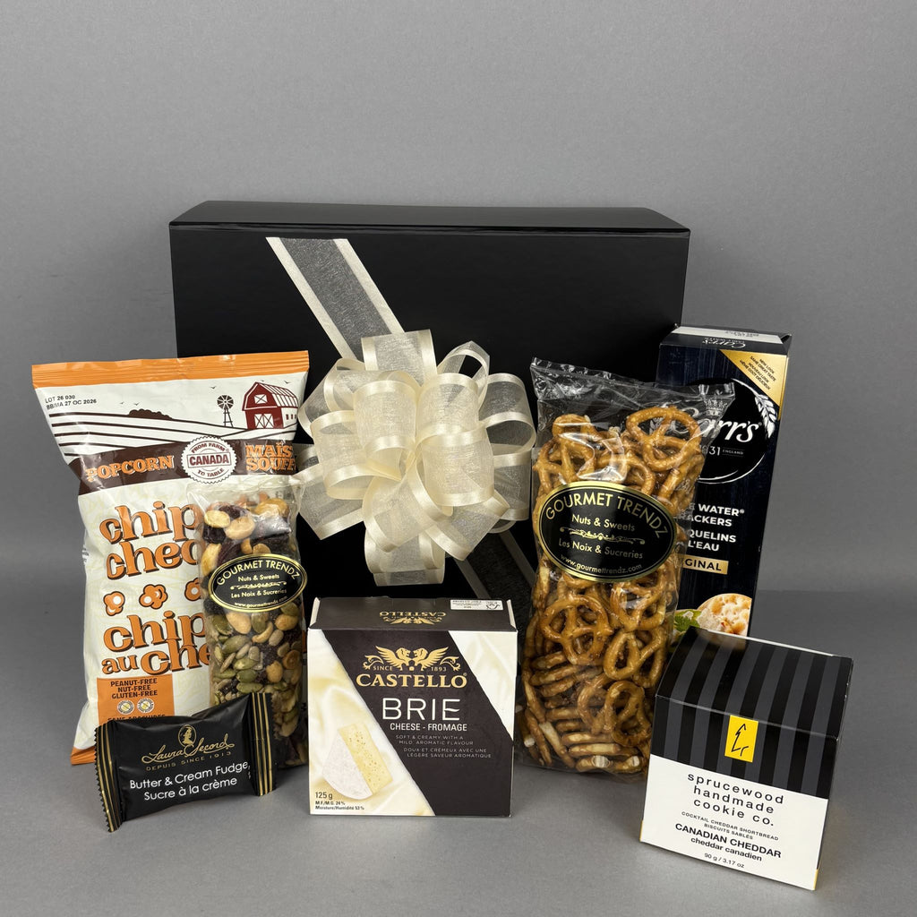 A gourmet gift hamper showcasing the black presentation box with a white ribbon, assorted snacks including popcorn, brie cheese, pretzels, and biscuits.