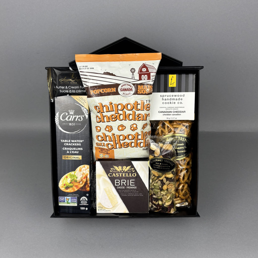 A curated gourmet food gift basket in a black magnet box, containing a Carr's water crackers, Laura Secord Butter & Cream Fudge, Chipotle Cheddar Popcorn, a box of Canadian Cheddar Biscuits, a box of Castello Brie cheese, a bag of pretzels, and mixed nuts.