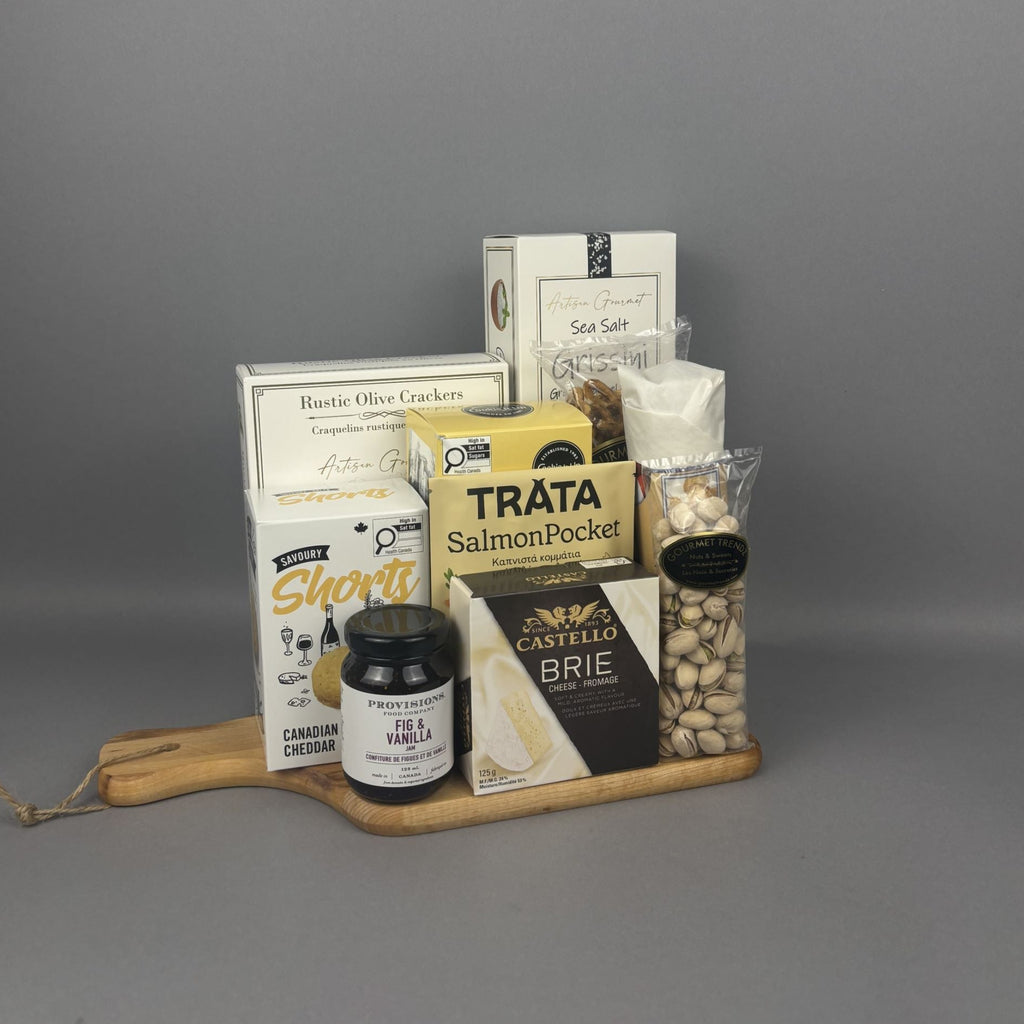 A curated gourmet gift basket arranged on a light wood serving board. The basket contains a variety of packaged food items including a box of Castello Brie cheese, a package of Tráta Smoked Salmon, a bag of Pistachios, a box of Rustic Olive Crackers, and a box of Sea Salt breadsticks.