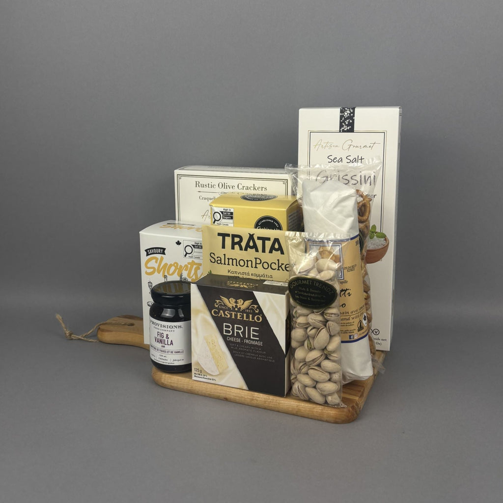 A gourmet gift hamper arranged on a small wooden tray. The hamper includes a box of rustic olive crackers, a package of Smoked Salmon, a box of Castello Brie cheese, a jar of Provisions fig & vanilla jam, a bag of pistachios, and dried salami stick.
