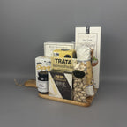 A gourmet gift hamper arranged on a small wooden tray. The hamper includes a box of rustic olive crackers, a package of Smoked Salmon, a box of Castello Brie cheese, a jar of Provisions fig & vanilla jam, a bag of pistachios, and dried salami stick.