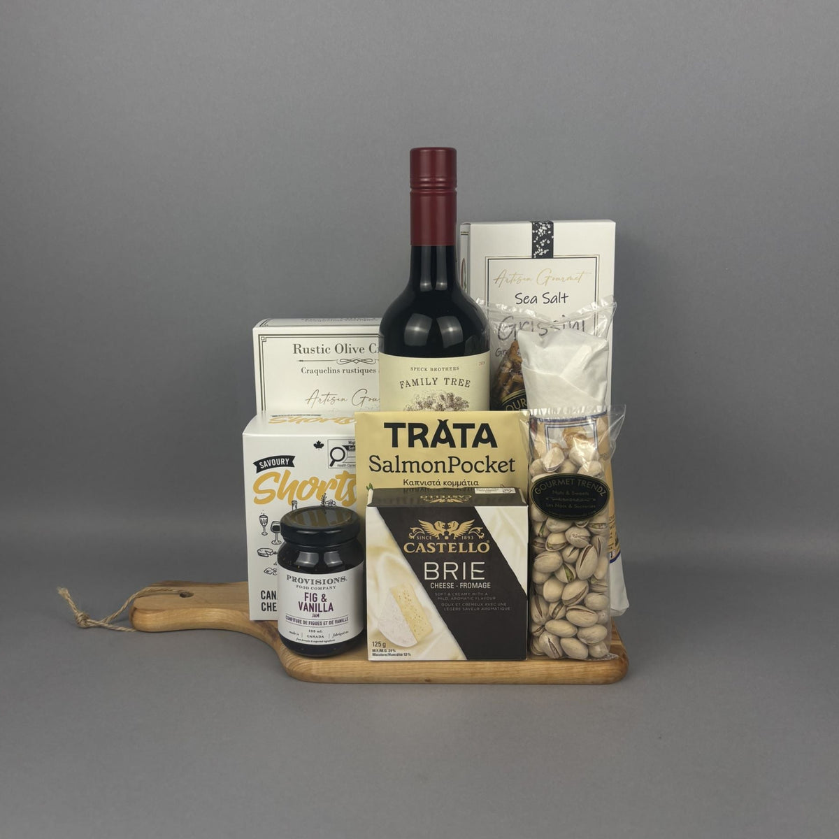 A gourmet gift hamper arranged on a wooden serving board. The hamper includes a bottle of red wine, a box of rustic olive crackers, a jar of Provisions jam, a box of Canadian Cheddar biscuits, a packet of Trâta Smoked Salmon, and a bag of pistachios, along with a box of Castello Brie cheese.