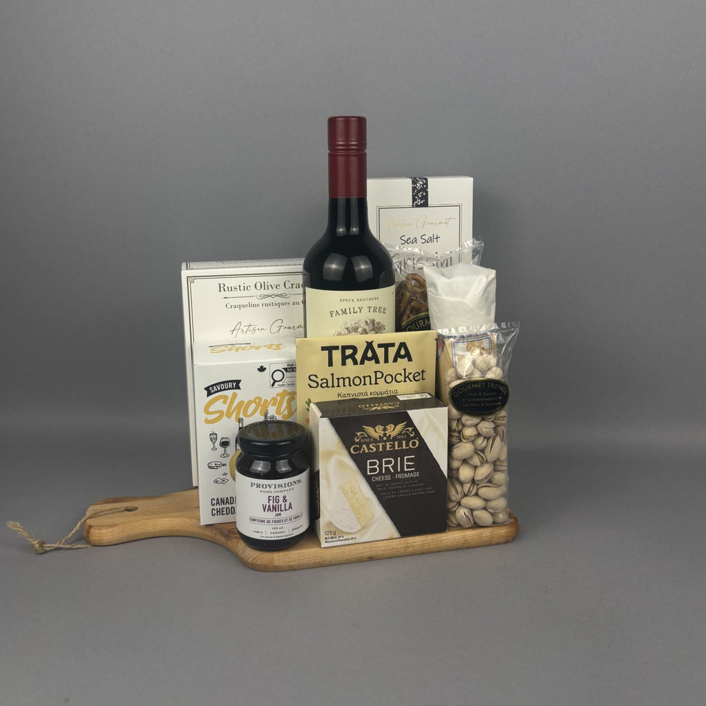A gourmet gift hamper arranged on a wooden serving board. The hamper includes a bottle of red wine, a box of rustic olive crackers, a jar of Provisions jam, a packet of smoked salmon, a box of Castello Brie, and a bag of pistachios.