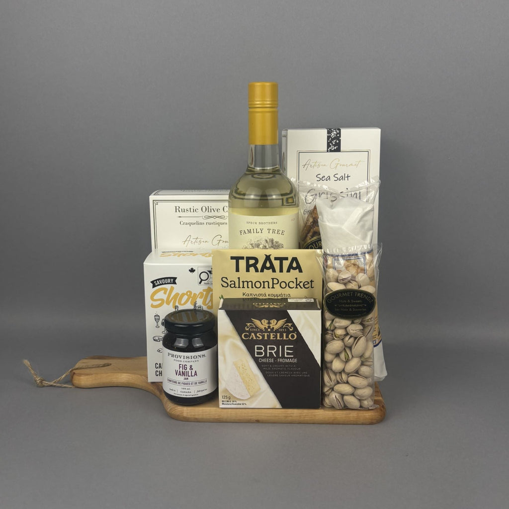 A gourmet gift hamper arranged on a small wooden tray. The set includes a package Trâta Smoked Salmon, a box of Castello Brie cheese, a jar of fig & vanilla jam, a box of Sea Salt breadsticks, and a bag of pistachios.