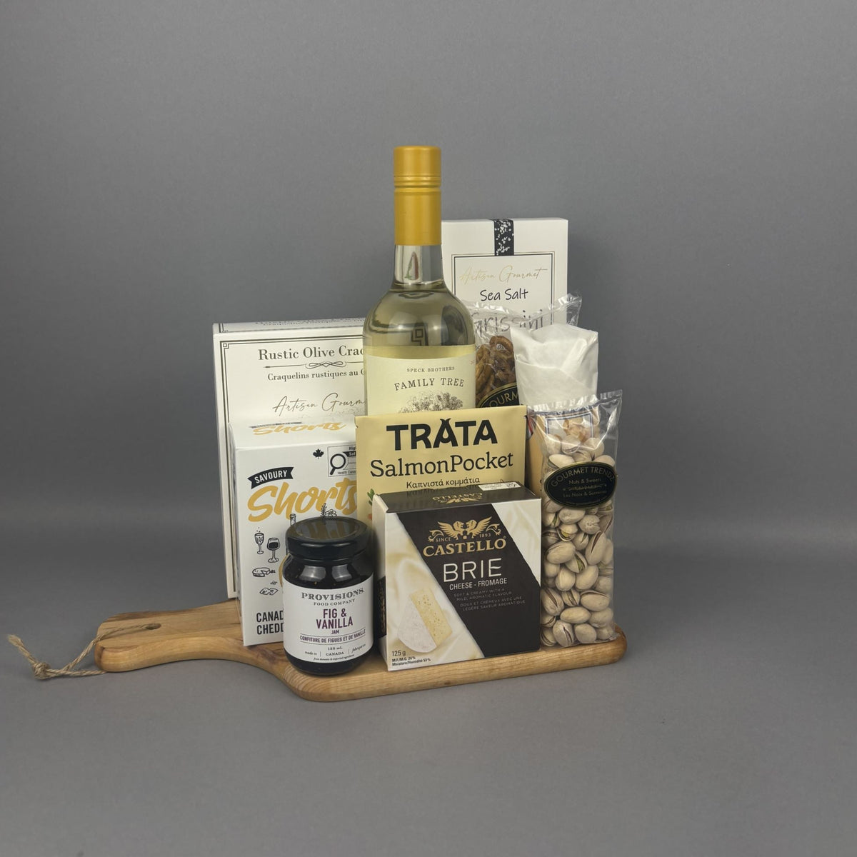 A gourmet gift hamper arranged on a small wooden serving board. The set includes a bottle of white wine, a box of rustic olive crackers, a package of Trâta Smoked Salmon, a box of Castello Brie cheese, a jar of Provisions jam, a bag of pistachios, and a dried salami stick.