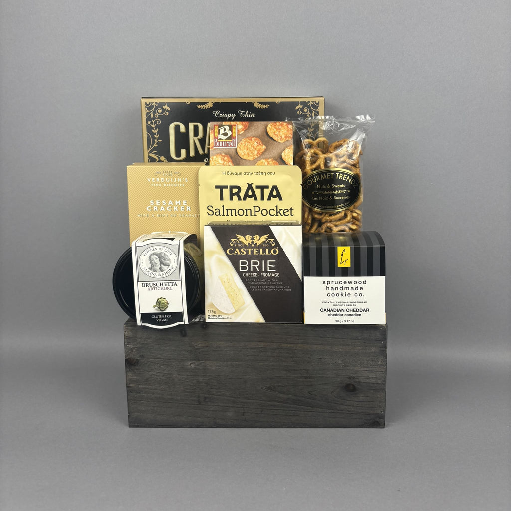 A gourmet gift hamper arranged in a dark wooden box. The hamper contains a box of artichoke bruschetta, a salmon pocket, a bag of gourmet pretzels, a Canadian Cheddar biscuits, and a box of crispy crackers.