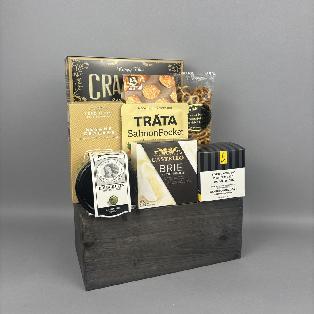 A gourmet gift hamper featuring an assortment of snacks and cheeses arranged in a dark wood crate. The hamper includes boxes of assorted crackers and biscuits, a bag of Tráta Smoked Salmon, a box of Castello Brie cheese, a bag of pretzels, and a jar of artichoke bruschetta.