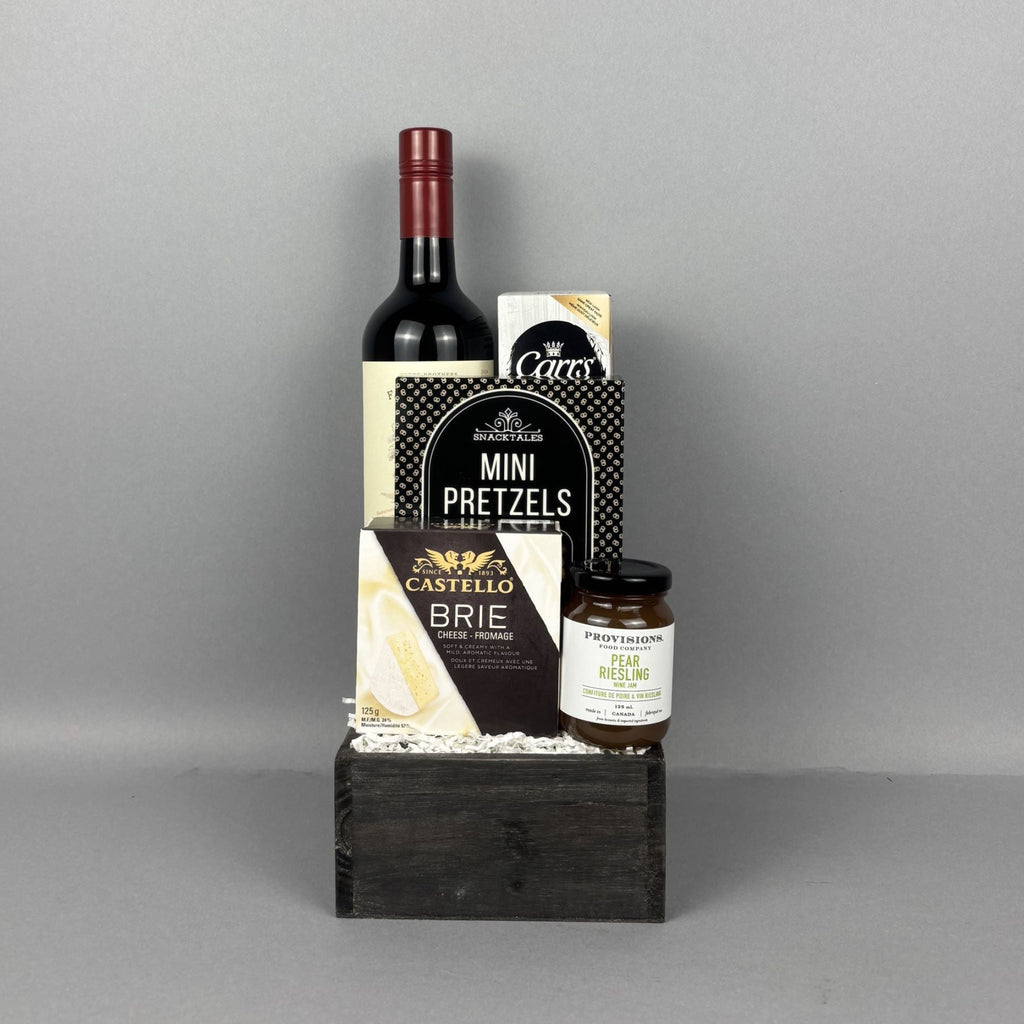 A gourmet gift hamper arranged on a dark wooden box. The set includes a bottle of red wine, Castello brie cheese, Mini Pretzels,, and a jar of Provisions pear riesling jam.