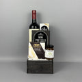 A gourmet gift hamper arranged on a dark wooden box. The set includes a bottle of red wine, Castello brie cheese, Mini Pretzels,, and a jar of Provisions pear riesling jam.