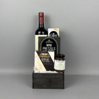 A gourmet gift hamper arranged on a dark wooden box. The set includes a bottle of red wine, Castello brie cheese, Mini Pretzels,, and a jar of Provisions pear riesling jam.
