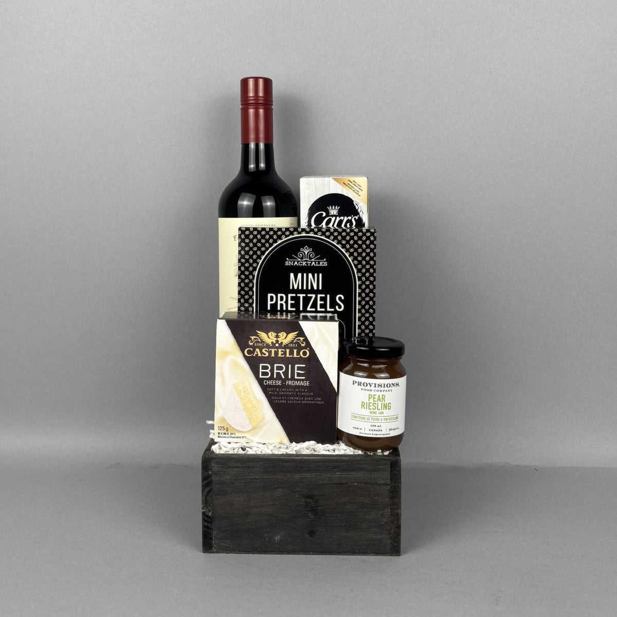 A gourmet gift hamper arranged on a dark wooden box. The set includes a bottle of red wine, Castello brie cheese, Mini Pretzels,, and a jar of Provisions pear riesling jam.