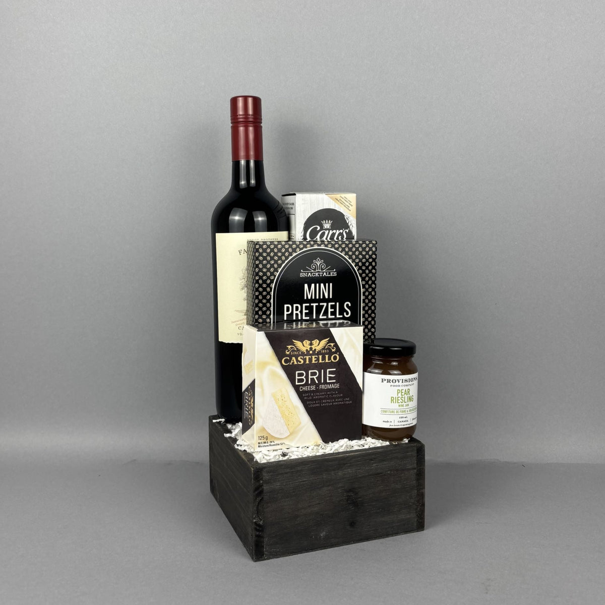 A gourmet gift basket in a dark wooden crate containing a bottle of red wine, a box of Mini Pretzels, a box of Castello Brie cheese, and a small jar of Provisions pear riesling jam, and Carr's black pepper crackers.