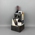 A gourmet gift basket arranged in a dark wooden crate. The basket contains a bottle of red wine, a box of Mini Pretzels, a box of Castello Brie cheese, a jar of Provisions jam, and a box of Carr's crackers.