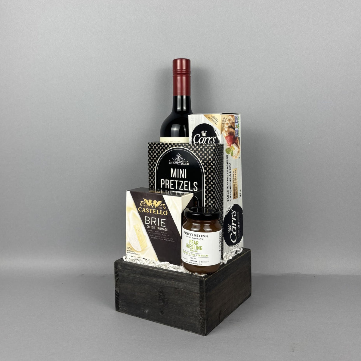 A gourmet gift basket arranged in a dark wooden crate. The basket contains a bottle of red wine, a box of Mini Pretzels, a box of Castello Brie cheese, a jar of Provisions jam, and a box of Carr's crackers.