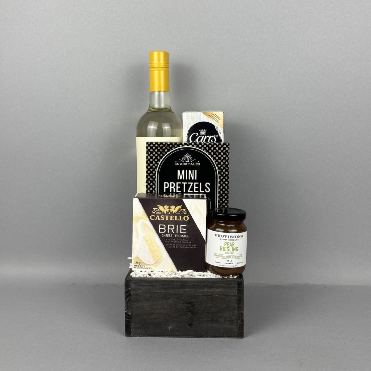 A gourmet gift basket set in a dark wooden crate, featuring a bottle of white wine, Mini Pretzels, Castello Brie cheese, and a jar of Provisions jam, and crackers. 