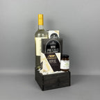 A gourmet gift basket set in a dark wooden crate, featuring a bottle of white wine, a box of Pretzels, a box of Castello Brie cheese, crackers, and a jar of pear riesling jam.