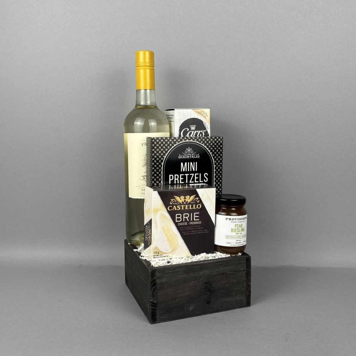 A gourmet gift basket set in a dark wooden crate, featuring a bottle of white wine, a box of Pretzels, a box of Castello Brie cheese, crackers, and a jar of pear riesling jam.
