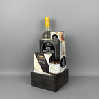 A gourmet gift basket set in a dark wooden crate, featuring a bottle of white wine, gourmet pretzels, brie cheese, and a small jar of Provisions jam.