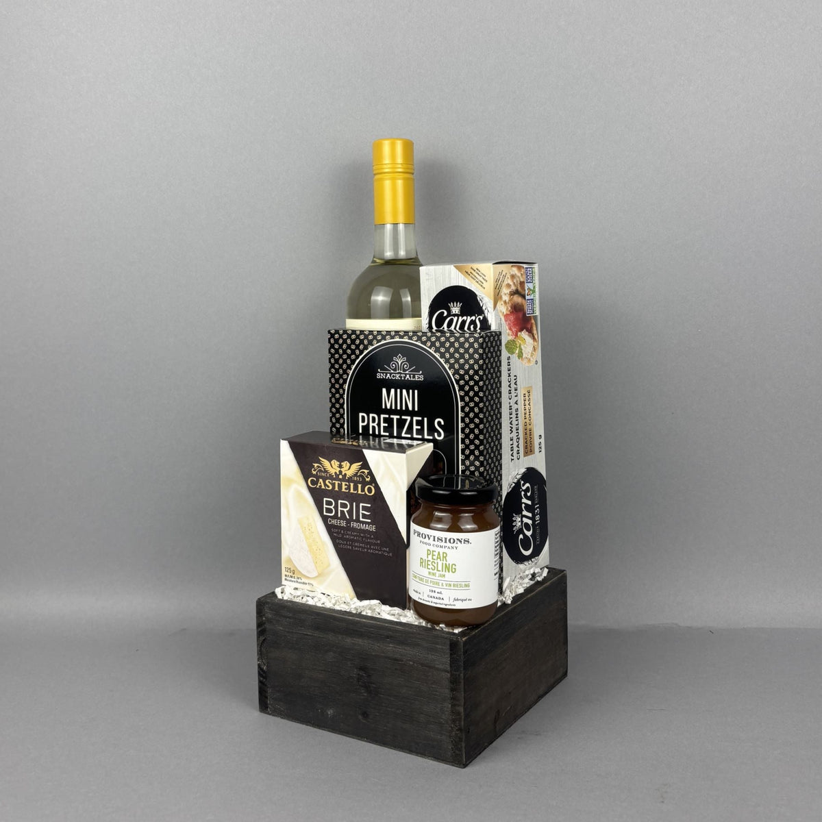 A gourmet gift basket set in a dark wooden crate, featuring a bottle of white wine, gourmet pretzels, brie cheese, and a small jar of Provisions jam.