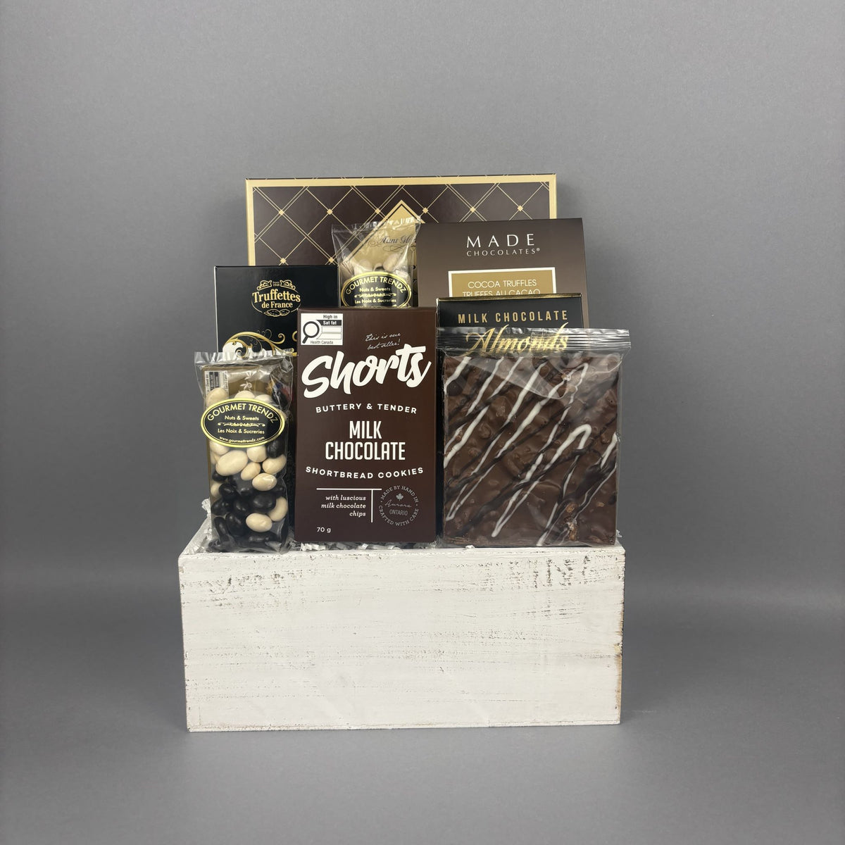 A chocolate gift basket arranged in a white wooden crate. The basket contains a box of Cookie It Up Milk Chocolate shortbread cookies, a box of Made Chocolate Toffee truffles, Chocolate Almonds, and a handcrafted bark bar.