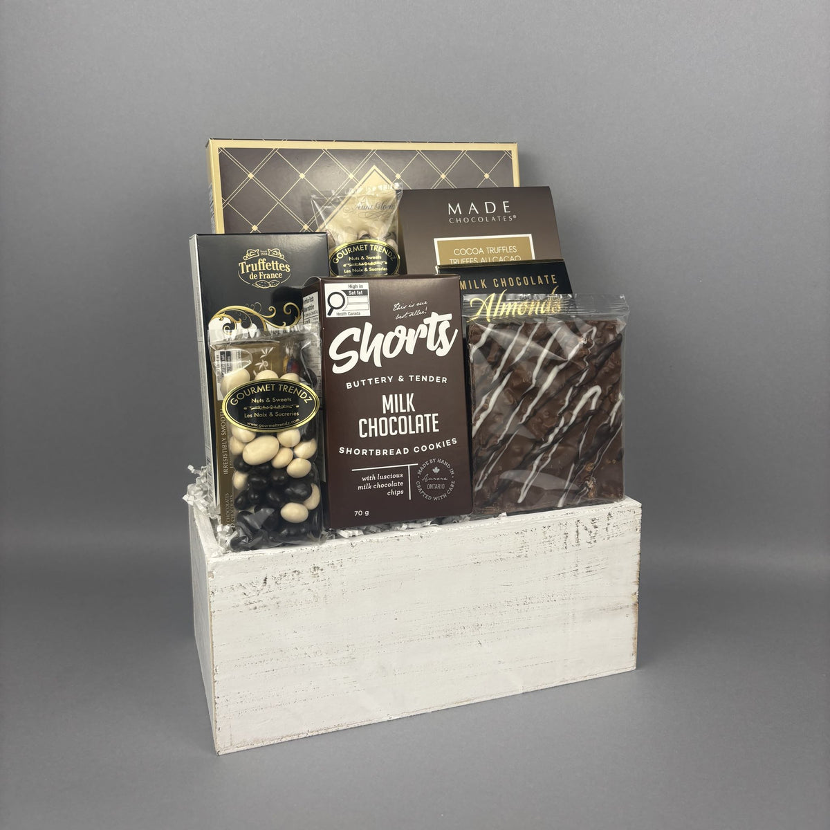 A gourmet chocolate gift basket arranged in a distressed white wooden box. The assortment includes Cookie It Up Chocolate Shortbread Cookies, Made Toffee Chocolate truffles, Milk Chocolate almonds, and Chocolate covered raisins.