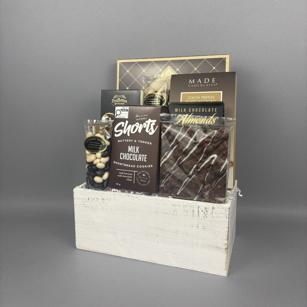 A chocolate gift basket arranged in a white wooden box. The basket contains assorted chocolates, including a milk chocolate shortbread cookies, a chocolate bark bar, a box of chocolate-covered nuts, and a decadent chocolate truffles.