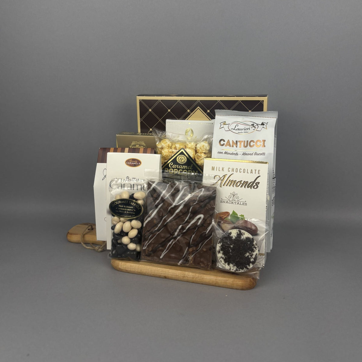 A gourmet chocolate and almond gift basket arranged on a wooden tray. The assortment includes a bag of Cantucci, milk chocolate almonds, a bag of gourmet caramel popcorn, a box of caramel candies, and a handcrafted chocolate bark bar.
