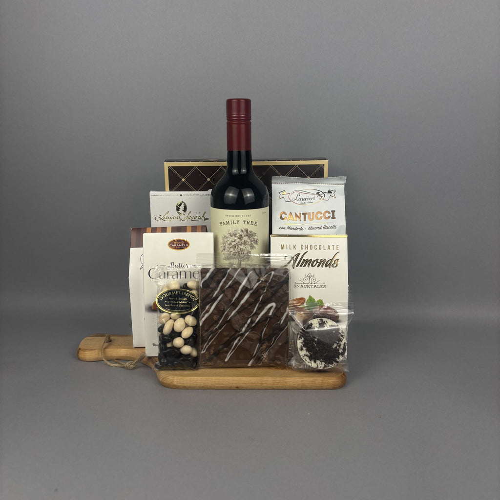 A gourmet gift hamper featuring a bottle of red wine, assorted chocolates, caramel candies, and almond chocolate on a wooden tray.