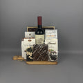 A gourmet gift hamper featuring a bottle of red wine, assorted chocolates, caramel candies, and almond chocolate on a wooden tray.