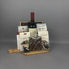 A gourmet gift hamper arranged on a wooden serving board. The hamper includes a bottle of red wine, assorted chocolate bars, milk chocolate almonds, butter caramels, and a sugar cookie box.
