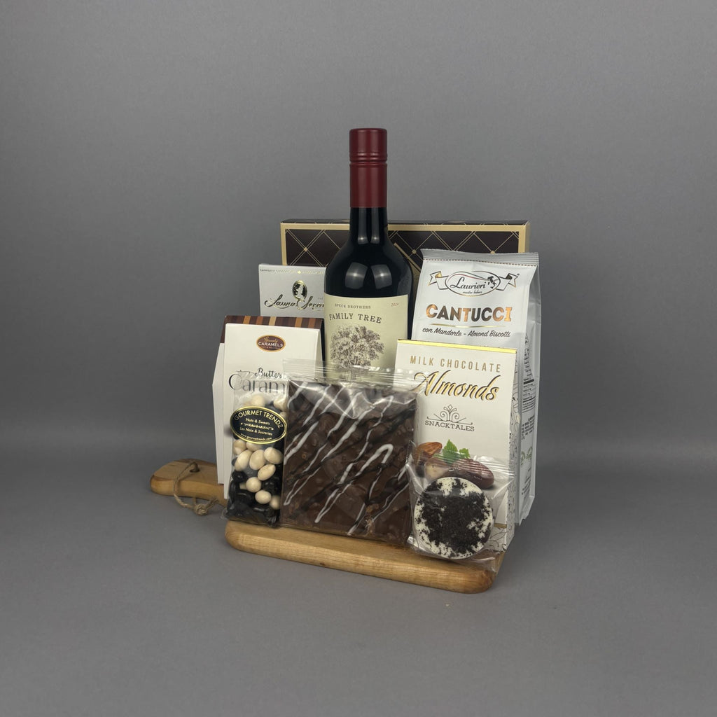 A gourmet gift basket arranged on a wooden tray. The basket includes a bottle of red wine, a bag of Cantucci, chocolate, almonds, sugar cookies, and assorted chocolates and nuts.