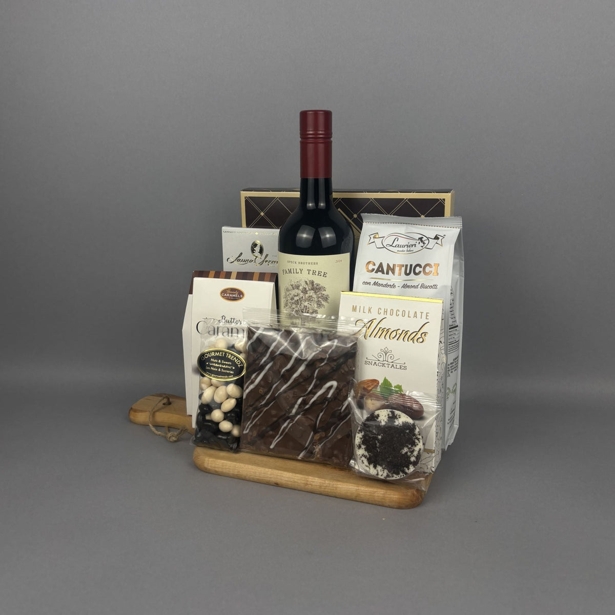 A gourmet gift basket arranged on a wooden tray. The basket includes a bottle of red wine, a bag of Cantucci, chocolate, almonds, sugar cookies, and assorted chocolates and nuts.