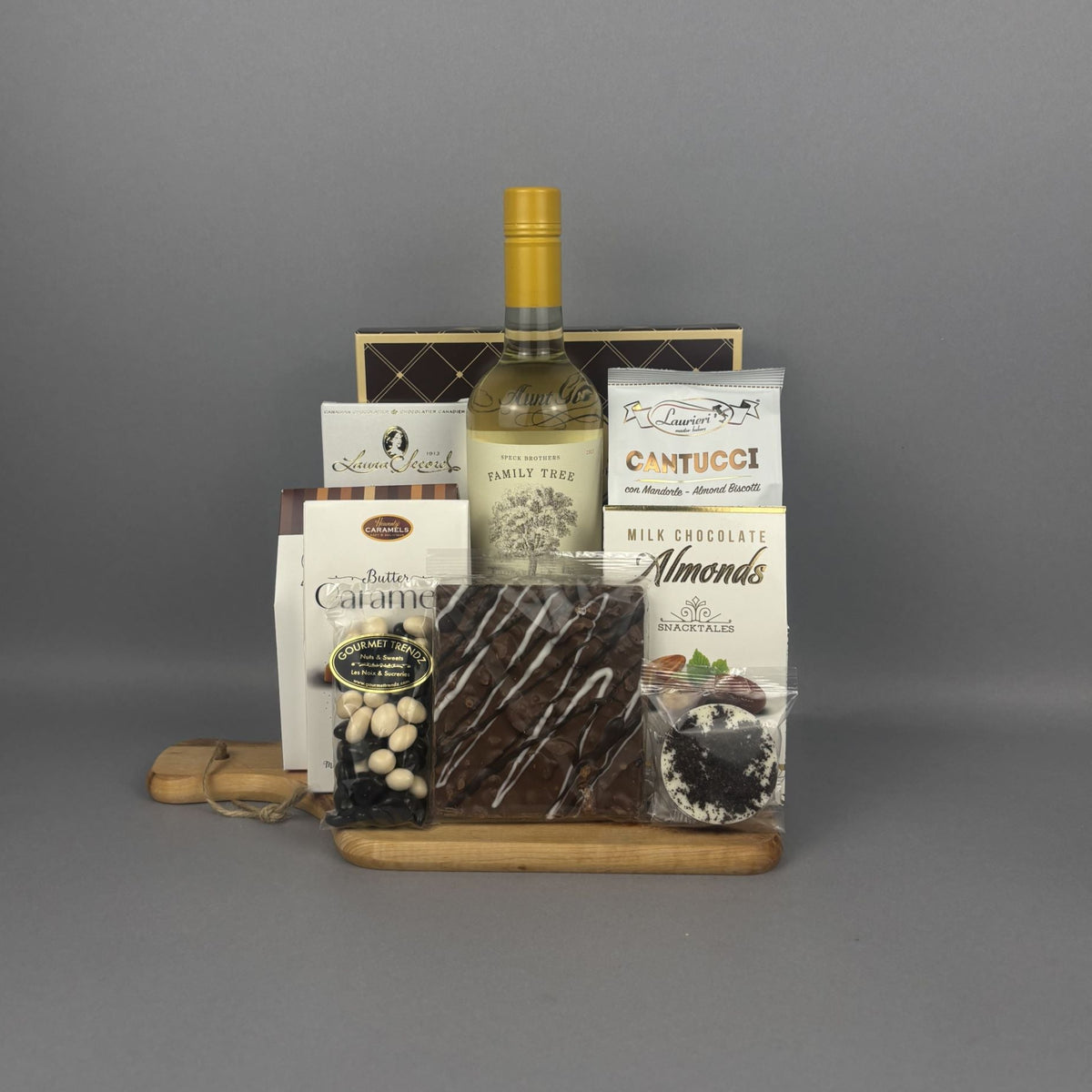 A gourmet gift hamper arranged on a small wooden tray. The hamper includes a bottle of white wine, a box of Cantucci, chocolate almonds, a bag of caramel candies, a bag chocolate-covered raisins, and other packaged treats.