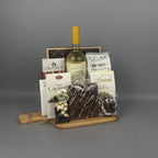 A gourmet gift hamper arranged on a wooden serving board. The hamper includes a bottle of white wine, assorted chocolate bars, a box of butter caramels,  chocolate almonds, a bag of almond cookies, and a bag of chocolate-covered raisins.