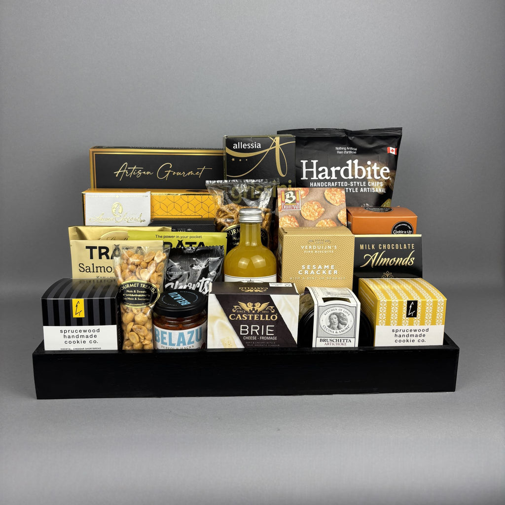 A gourmet gift hamper arranged on a black tray, featuring a large assortment of snacks, cheeses, crackers, and a small bottle of mandarin vinegar.