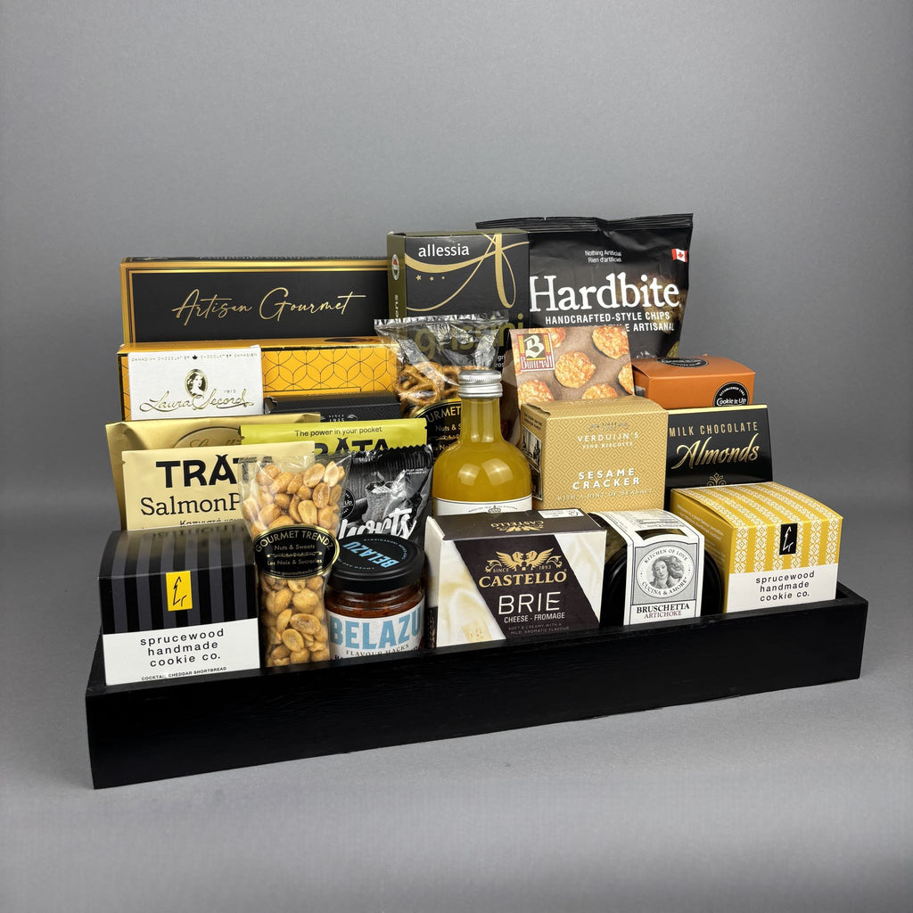 A gourmet gift hamper arranged on a black rectangular tray. The assortment includes packaged snacks, cheeses, crackers, nuts, and a small bottle of vinega, with visible brands such as Hardbite, Castello, Belazu, and Laura Secord.