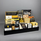 A gourmet gift hamper arranged on a black rectangular tray. The assortment includes packaged snacks, cheeses, crackers, nuts, and a small bottle of vinega, with visible brands such as Hardbite, Castello, Belazu, and Laura Secord.