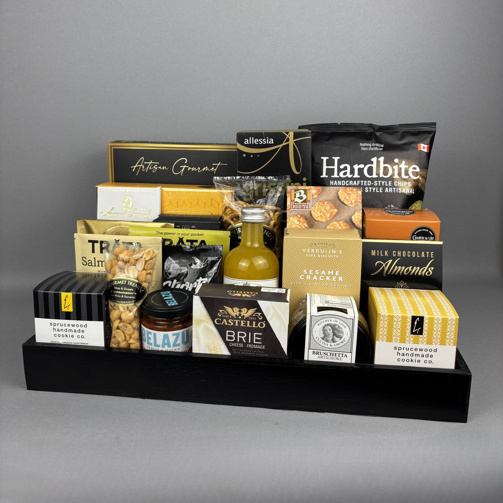 A gourmet food gift basket arranged on a black rectangular tray. The basket contains assorted packaged snacks and treats, including Hardbite chips, Castello Brie cheese, Hamlet chocolates, and several other boxed and bagged foods.