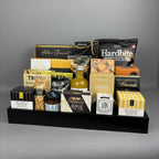 A gourmet food gift basket arranged on a black rectangular tray. The basket contains assorted packaged snacks and treats, including Hardbite chips, Castello Brie cheese, Hamlet chocolates, and several other boxed and bagged foods.