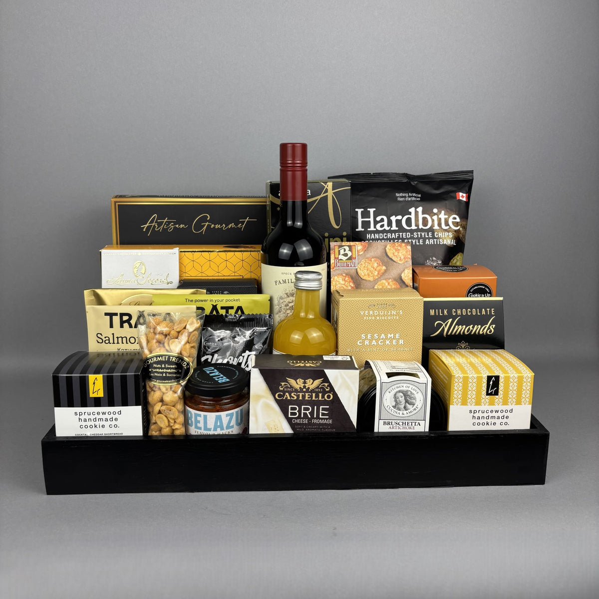A gourmet food gift hamper arranged on a black tray. The assortment includes a bottle of red wine, a small bottle of mandarin vinegar, bags of nuts and crackers, boxes of chocolate and cheese, and other packaged snacks.